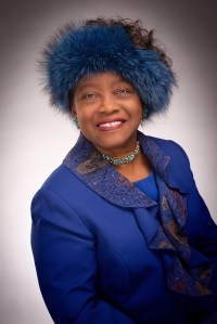 Lura Smith smiling and posed in a royal blue blazer and blue feathered headpiece. 